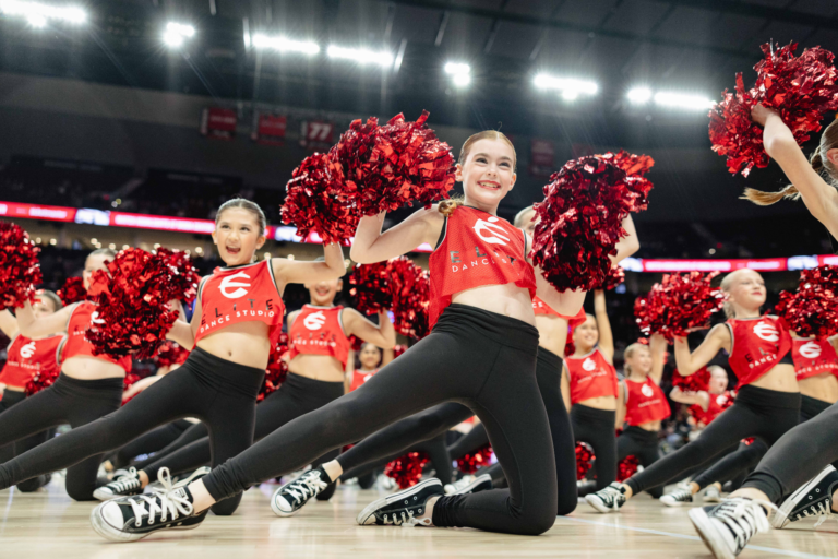 Young dancers in red uniforms performing a cheer routine with pom-poms on a brightly lit stage.