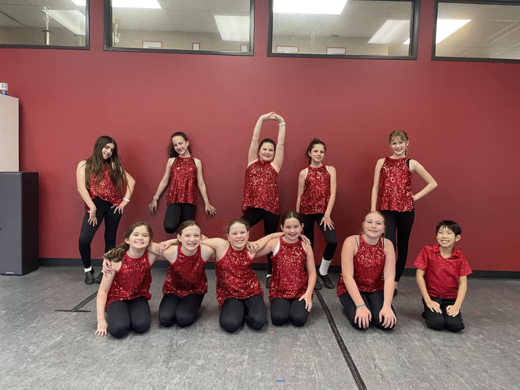 Group of young dancers in red sequin tops and black pants posing together in a studio with red walls.